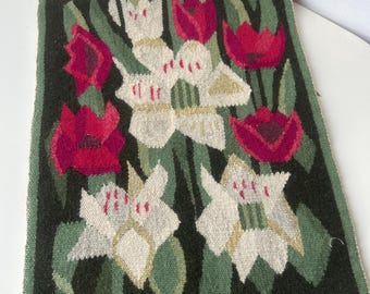 Op de afbeelding: Een geweven wandtapijt met een bloemenpatroon. Het wandtapijt toont witte en rode bloemen met groene bladeren op een donkergroene achtergrond. De bloemen zijn gestileerd, met gedetailleerde bloemblaadjes en stelen.