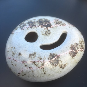 May include: A white ceramic stone with a brown and pink speckled glaze. The stone has a cut-out smiley face design.