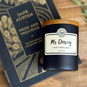 May include: Black candle jar with a wooden lid and a white label that reads "Mr. Darcy" with the text "musk, vanilla, spices" and "hand-poured" below. The candle is sitting on top of a book with a gold and black cover that reads "Jane Austen, Pride and Prejudice".