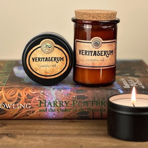 May include: Two candles with black lids and labels that say "Veritaserum - vanilla - oud" on top of a Harry Potter book. The book is titled "Harry Potter and the Order of the Phoenix" by J.K. Rowling.