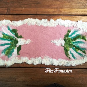 Könnte beinhalten: Ein rosa Filzteppich mit weißem Rand und zwei grünen und weißen Blumendesigns. Der Teppich trägt den Text "Filz Fantasien" in der Mitte unten.