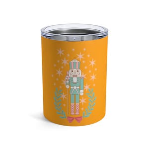 Puede incluir: Un vaso amarillo con un diseño festivo de cascanueces. La ilustración incluye un soldado cascanueces en verde azulado y rosa, rodeado de estrellas y una corona de laurel. El vaso tiene un borde plateado y un interior negro.