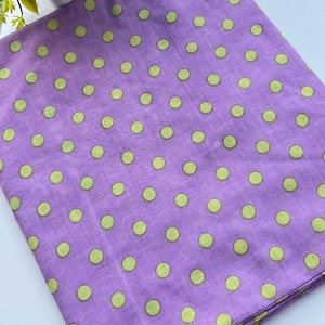 Puede incluir: Una pieza de tela doblada de color morado claro que presenta un patrón repetitivo de pequeños lunares de color verde lima sólido dispuestos en una cuadrícula en toda la superficie del material textil, mostrando un diseño consistente.