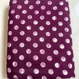 May include: A folded fabric with a deep purple background and a repeating pattern of large, white and pink moon-like circles. The fabric appears to be cotton or a similar material, suitable for sewing projects.