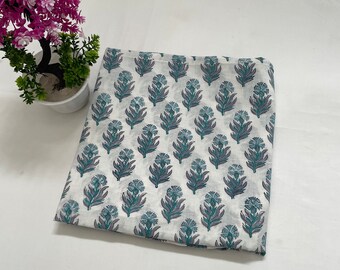 May include: White fabric with a repeating floral pattern in teal and gray. The flowers have a stylized design. The fabric is folded and appears to be cotton or a similar material. A small potted plant with pink flowers is in the background.