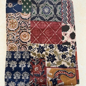 Op de afbeelding: Een gevouwen textiel met een patchwork ontwerp. De stof heeft een verscheidenheid aan patronen, waaronder bloemen, geometrische en paisley motieven. Het kleurenpalet omvat marine, bordeauxrood, bruin en crème.