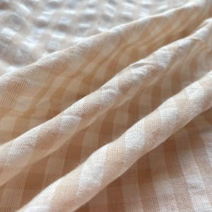 May include: A lightweight fabric featuring a soft beige and white gingham check pattern. The material is draped in gentle folds, highlighting the woven texture of the textile and the consistent grid design across the surface of the cloth.