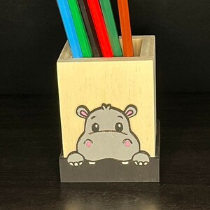 May include: A wooden pencil holder with a cute cartoon hippopotamus peeking out from the top. The holder is painted in natural wood tones and has a black base. It is filled with colorful pencils.