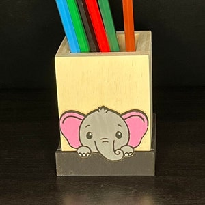 Peut inclure: Un porte-crayons en bois avec un adorable éléphant de dessin animé qui sort du haut. Le porte-crayons est peint dans des tons de bois naturels et a une base noire. Le porte-crayons est rempli de crayons de couleur.
