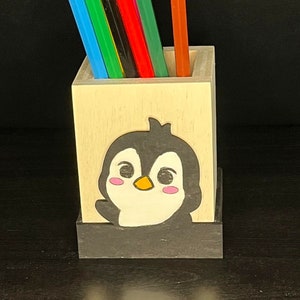 Puede incluir: Un portalápices de madera con diseño de pingüino. El soporte es de color claro con una base negra y presenta un pingüino de dibujos animados con mejillas rosadas y pico amarillo. Contiene lápices de colores.