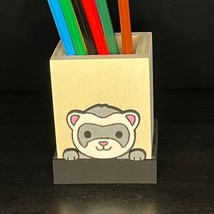Peut inclure: Un porte-crayons en bois avec un furet de dessin animé qui sort du haut. Le porte-crayons est peint dans une couleur bois clair et a une base noire. Le furet est gris avec des oreilles roses et un nez blanc.
