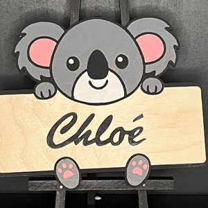 May include: A wooden sign with a grey koala holding the sign. The sign says "Chloé" in black cursive writing.