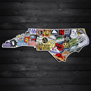 May include: A white outline of the state of North Carolina, filled with colorful brewery logos and stickers. The logos feature various designs and text, including "Hi-Wire Region" and "26". The background is a dark wood grain.
