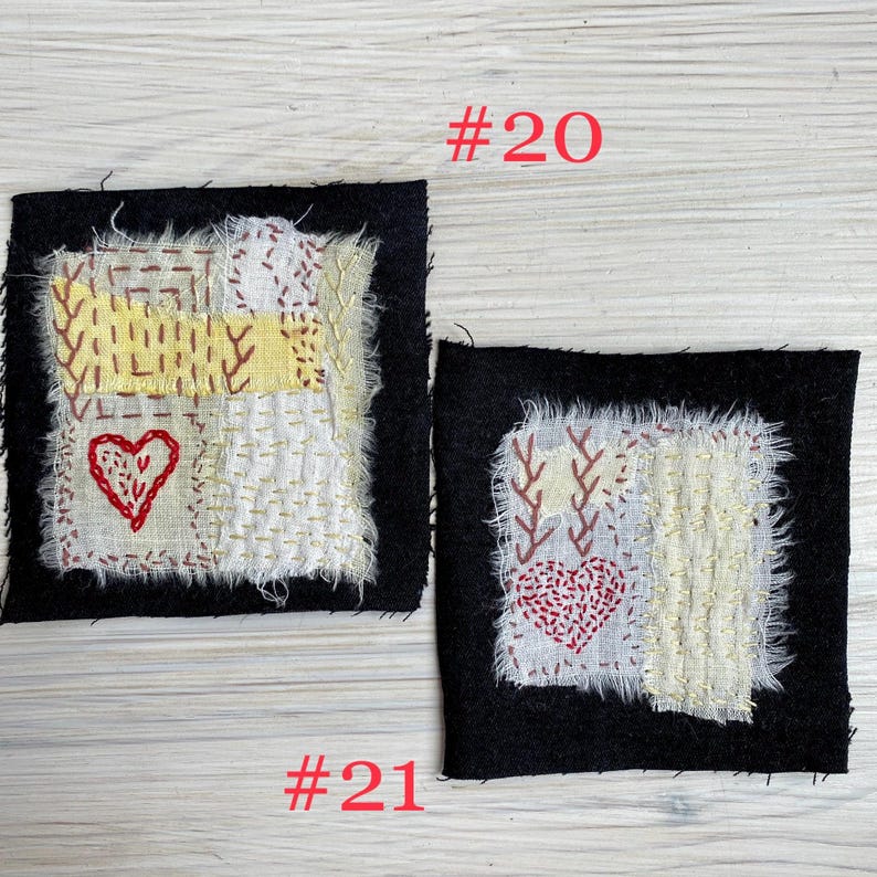 May include: Two square fabric patches labeled #20 and #21. Each patch features a black square base with a central white and yellow frayed fabric square, decorated with red embroidered hearts and intricate hand-stitched running thread patterns.