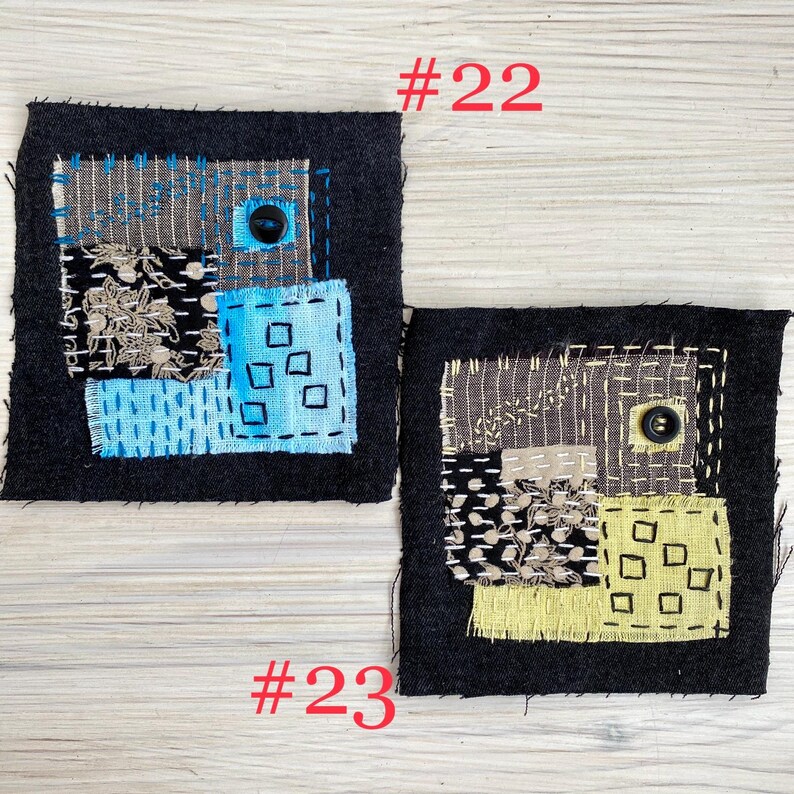 May include: Two square fabric patches featuring visible mending with sashiko-style stitching. Patch #22 has blue and black patterned fabric with a dark button. Patch #23 has yellow and black patterned fabric with a dark button and white stitching.