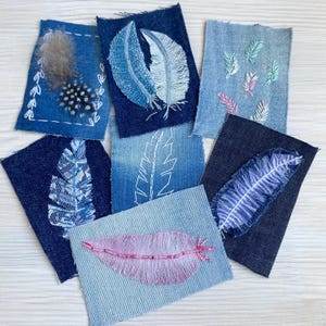 May include: A collection of seven rectangular denim fabric patches featuring various feather designs. The patches display embroidery, lace, beads, and real feathers in shades of blue, white, and pink, showcasing different textures and stitching techniques.