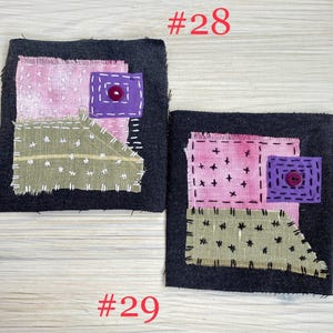May include: Two square dark gray fabric patches featuring layered textile appliqu&eacute;s. Each patch includes a pink square, a sage green irregular shape, and a small purple square with a centered button, all secured with visible white or black stitching.