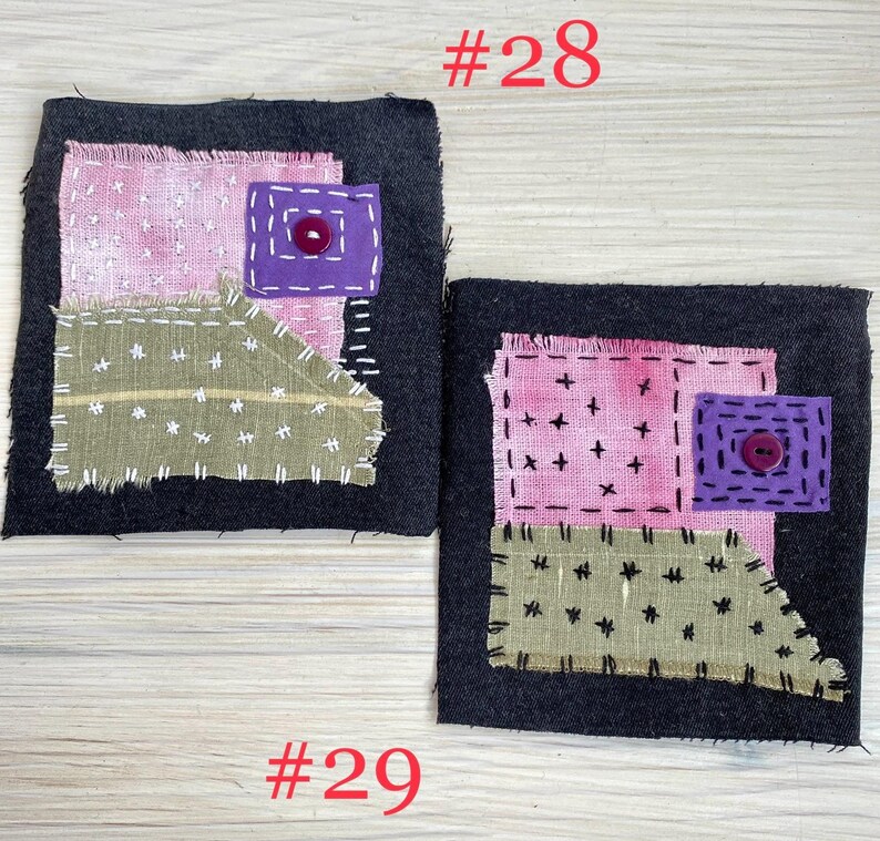 May include: Two square dark gray fabric patches featuring layered textile appliqu&eacute;s. Each patch includes a pink square, a sage green irregular shape, and a small purple square with a centered button, all secured with visible white or black stitching.