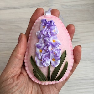 Puede incluir: Adorno de huevo de Pascua de fieltro rosa con un diseño de flor de cinta lavanda y blanca. La flor tiene detalles amarillos y hojas de cinta verde. Se adjunta un lazo de cinta blanca para colgar.