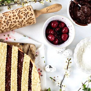 May include: A wooden rolling pin with a floral and bunny design, a white bowl with red cherries, a white bowl with chocolate filling, a white bowl with flour, and a branch of white blossoms. The rolling pin is labelled "Folkroll".