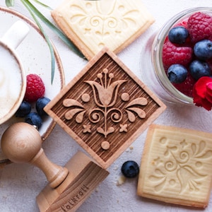 May include: A wooden cookie stamp with a floral design, a cup of coffee, and several cookies with the same floral design. The stamp is labeled "Folkrou" and is surrounded by red carnations, blueberries, and raspberries.