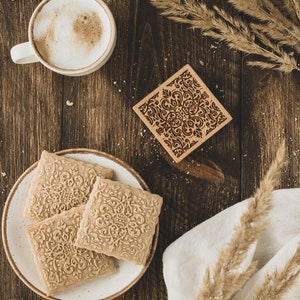 Op de afbeelding: Een houten tafel met een kop koffie, een bord met koekjes, een houten stempel en droog gras. De koekjes zijn versierd met een bloemmotief.