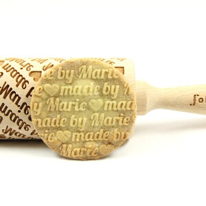 Puede incluir: Un rodillo de madera con un mango y el texto "folkroll" grabado en el mango. El rodillo está grabado con el texto repetido "made by Marie" y corazones. Una galleta con el mismo texto y corazones se muestra junto al rodillo.