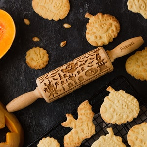 Può includere: Un mattarello di legno con un design intagliato a tema Halloween, tra cui pipistrelli, una casa infestata e lapidi, è posto tra biscotti a forma di zucca. Una zucca intagliata e una ciotola di polpa di zucca sono anch'esse nell'immagine.