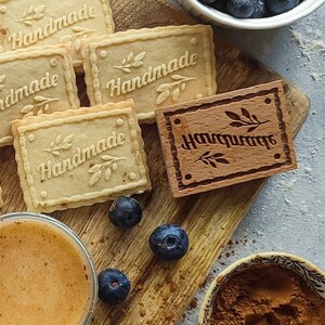 May include: A wooden cutting board with five homemade cookies and a wooden stamp that says "Handmade". The cookies are arranged in a circle around the stamp. There is a bowl of blueberries, a cup of cinnamon, and a glass of orange juice on the board.
