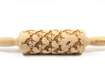 May include: A wooden rolling pin with a repeating pattern of cats chasing a mouse. The rolling pin is engraved with the word "J'oikroll".