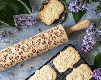 May include: A wooden rolling pin with a carved design of birds and flowers. The rolling pin is engraved with the text "Folkroll". The rolling pin is used to create decorative patterns on cookies. There are several cookies with the same design on a wire rack.