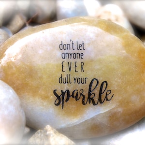 May include: A smooth, light brown stone with the inspirational message "don't let anyone EVER dull your sparkle" engraved in black lettering.
