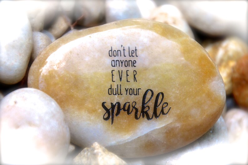 May include: A smooth, light brown stone with the inspirational message "don't let anyone EVER dull your sparkle" engraved in black lettering.