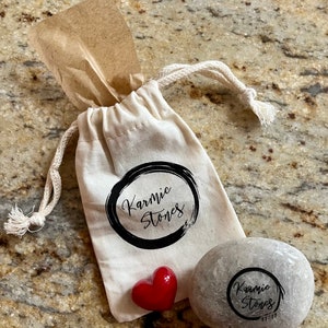 May include: A small, gray, round stone with the words "Karmic Stones" and "11-11" inscribed on it. The stone is in a white drawstring bag with the words "Karmic Stones" printed on it. A red heart-shaped stone is also in the bag.