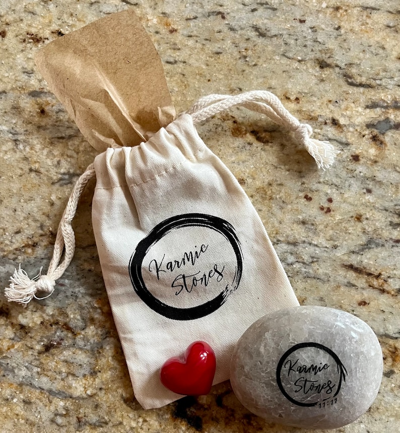 May include: A small, gray, round stone with the words "Karmic Stones" and "11-11" inscribed on it. The stone is in a white drawstring bag with the words "Karmic Stones" printed on it. A red heart-shaped stone is also in the bag.