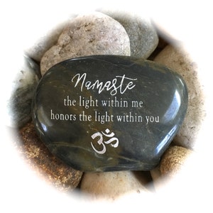 Op de afbeelding: Een donkergrijze steen met de woorden "Namaste the light within me honors the light within you" en het Om-symbool gegraveerd in zilver.