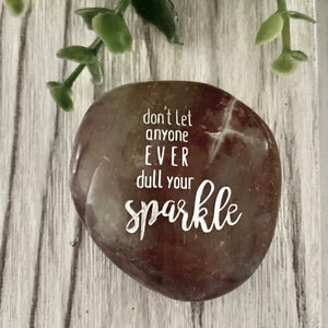 May include: A brown and white stone with the text "don't let anyone EVER dull your sparkle" engraved on it.