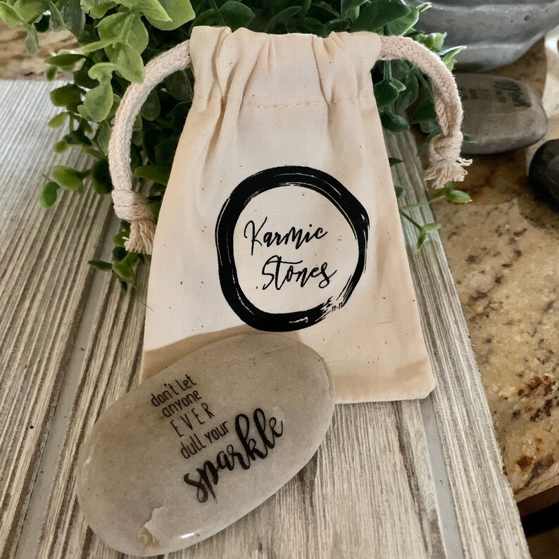 May include: A small, gray, oval-shaped stone with the text "don't let anyone EVER dull your sparkle" printed on it. The stone is in a white drawstring bag with the text "Karmic Stones" printed on it.
