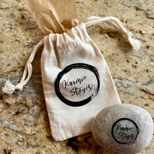 May include: A small, light gray stone with the words "Karmic Stones" written in black ink inside a circle. The stone is sitting next to a white drawstring bag with the same words and design printed on it.