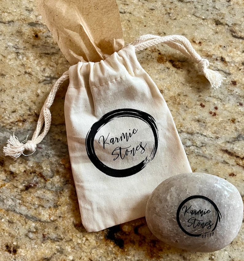 May include: A small, light gray stone with the words "Karmic Stones" written in black ink inside a circle. The stone is sitting next to a white drawstring bag with the same words and design printed on it.