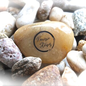 May include: A close-up of a smooth, light brown stone with the words "Karmic Stones" written in black ink inside a circle. The stone is surrounded by other stones of various colors and textures.