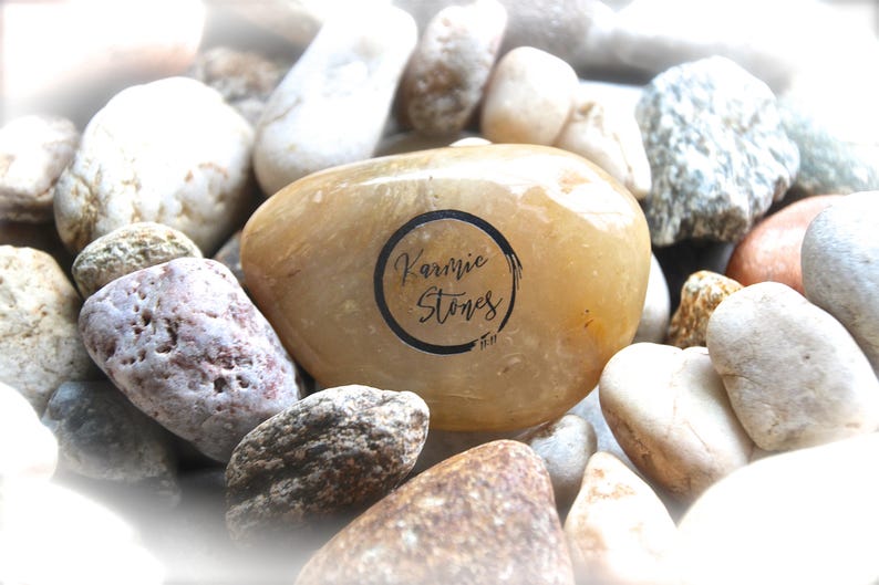 May include: A close-up of a smooth, light brown stone with the words "Karmic Stones" written in black ink inside a circle. The stone is surrounded by other stones of various colors and textures.
