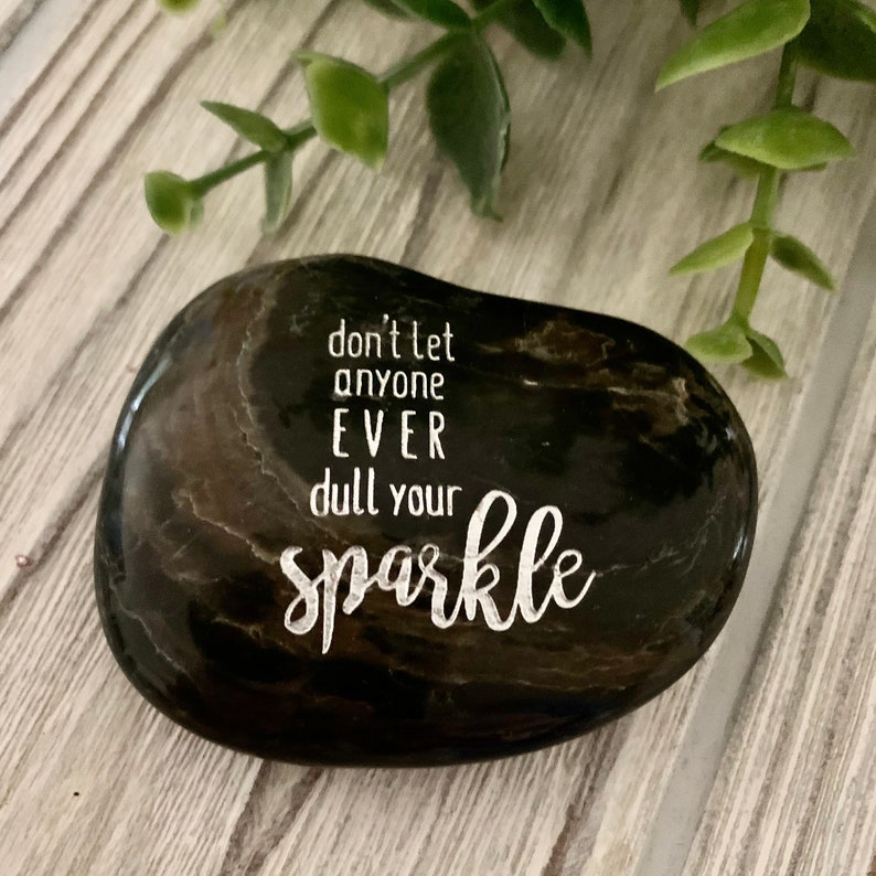 May include: A black and gray smooth river rock with the words "don't let anyone EVER dull your sparkle" engraved in white.