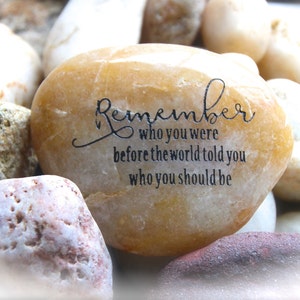 May include: A smooth, light brown stone with the inscription "Remember who you were before the world told you who you should be".