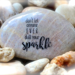 May include: A smooth, light brown stone with the inspirational message "don't let anyone EVER dull your sparkle" written in black ink.