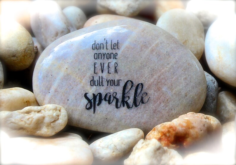 May include: A smooth, light brown stone with the inspirational message "don't let anyone EVER dull your sparkle" written in black ink.
