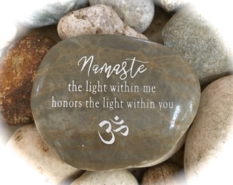 Op de afbeelding: Een grijze steen met de woorden "Namaste the light within me honors the light within you" en het symbool "Om" erop gegraveerd.