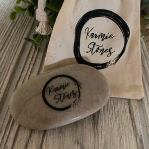 May include: A gray stone with the words "Karmic Stones" written in black ink inside a circle. The stone is sitting on a wooden surface next to a white cloth bag with the same logo.