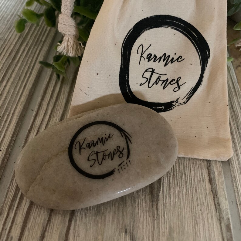 May include: A gray stone with the words "Karmic Stones" written in black ink inside a circle. The stone is sitting on a wooden surface next to a white cloth bag with the same logo.