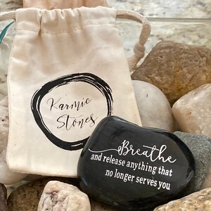 May include: A black smooth stone with the words "Breathe and release anything that no longer serves you" engraved on it. The stone is in a white drawstring bag with the words "Karmic Stones" printed on it.